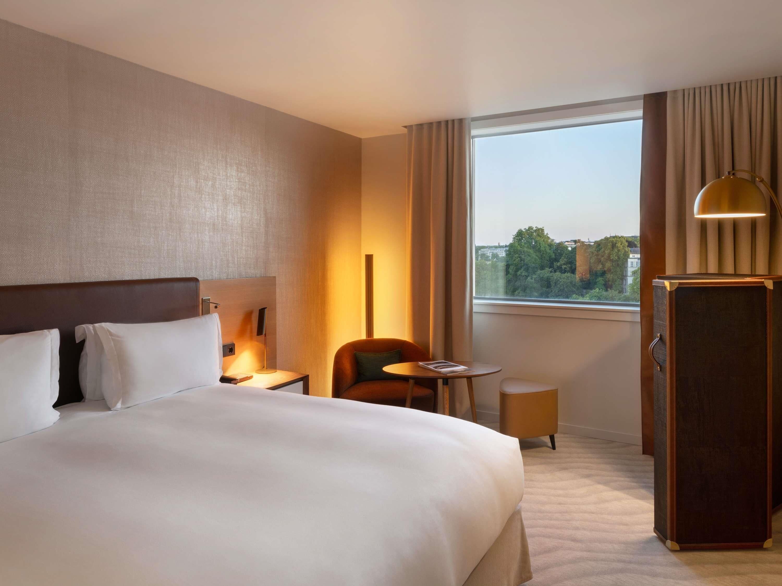 A hotel room with a large bed, a desk, a chair, a small table, a lamp, and a wardrobe.