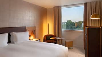 A hotel room with a large bed, a desk, a chair, a small table, a lamp, and a wardrobe.