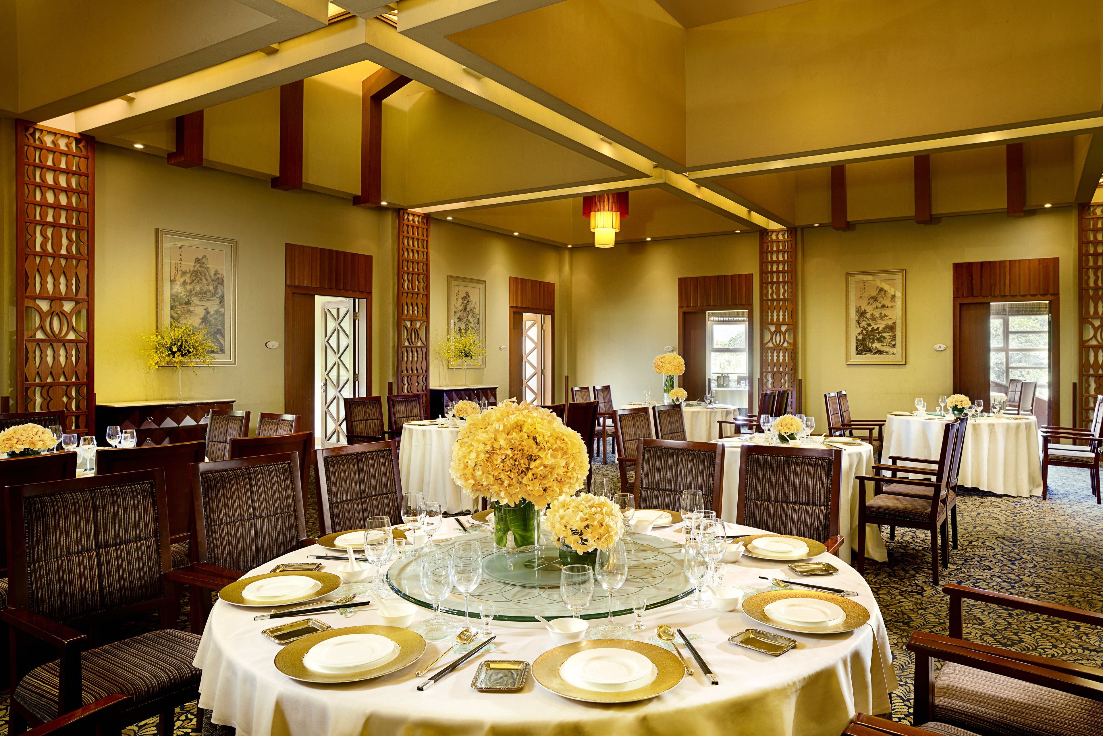 A formal dining room with round tables set for multiple guests, featuring gold-rimmed plates and clear glassware.