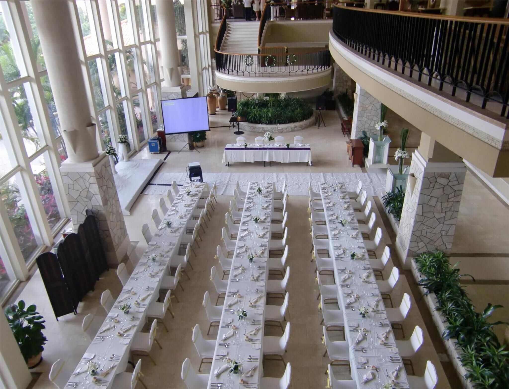 A spacious indoor venue with multiple long tables set for dining, a central stage, and a balcony overlooking the area.