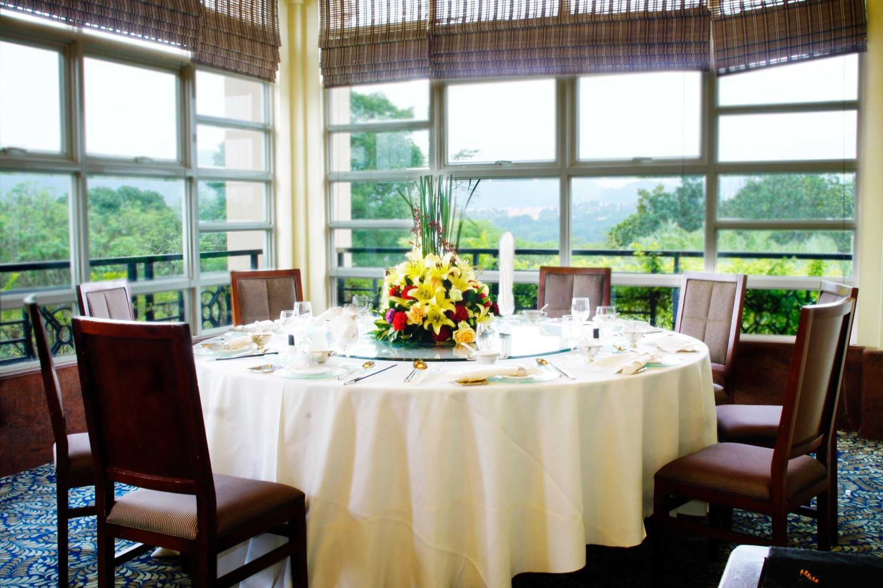 A round dining table set for four with a white tablecloth, surrounded by wooden chairs. A floral centerpiece is on the table, and the room has large windows with a view of greenery outside.
