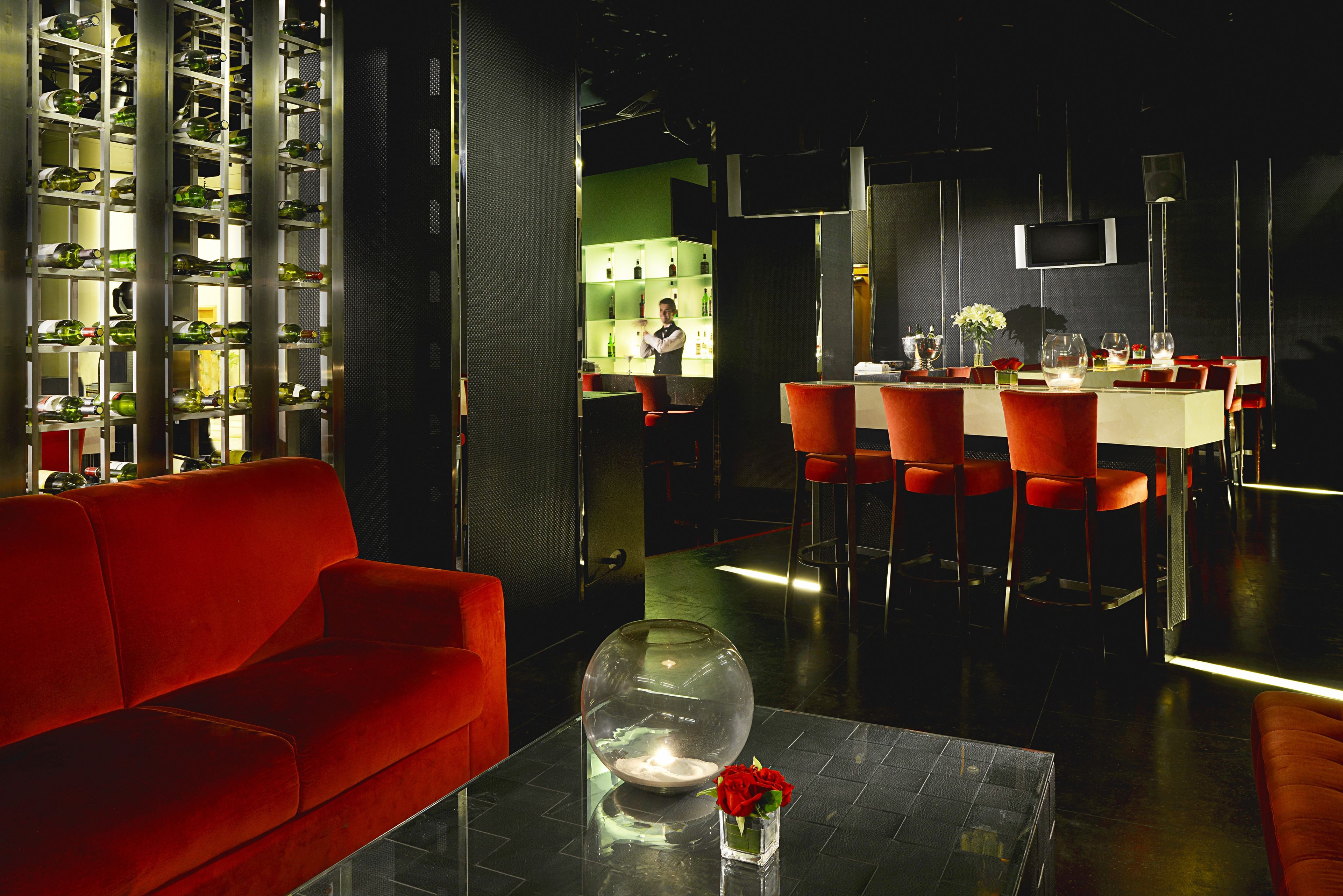 Un interior moderno con un sofá rojo, una mesa de centro de cristal con un jarrón de flores, y una zona de bar con sillas rojas y un cantinero.
