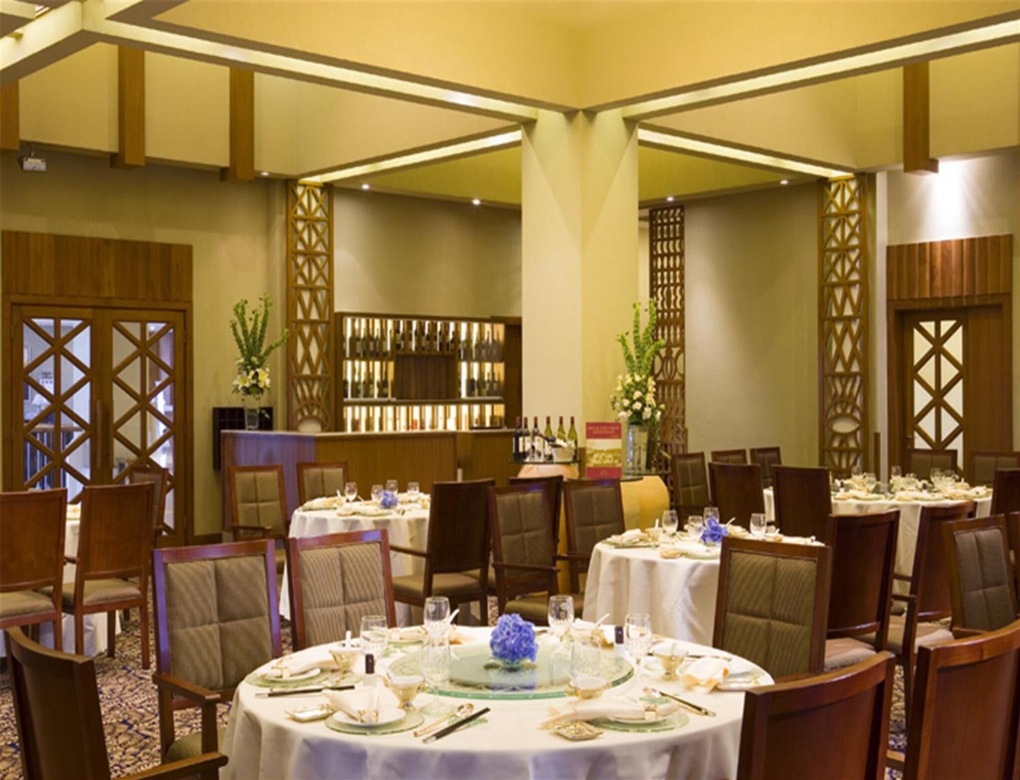 A banquet hall with round tables set for dining, a bar area with bottles, and decorative plants.