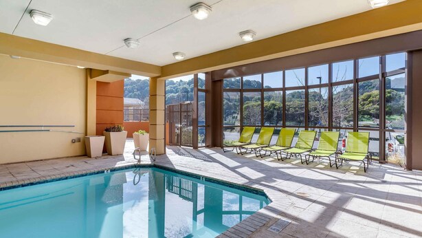 A swimming pool with clear blue water, surrounded by a tiled floor and beige walls. There are yellow chairs and a small planter with plants near the pool.