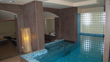 Una piscina interior moderna con suelo y paredes de azulejos, una zona de estar con sillones, y un pilar con una lámpara.