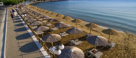 A beach with rows of sun loungers and thatched umbrellas, a paved walkway, and a mountain in the background.