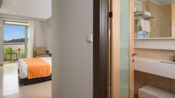A hotel room with a bed, a bathroom with a sink and mirror, and a balcony with a chair and table. - Corissia Princess Hotel (Apokoronas)