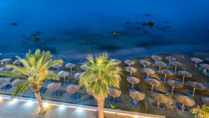 A beachside view with rows of sun loungers and umbrellas, palm trees, and a calm sea. - Corissia Princess Hotel (Apokoronas)