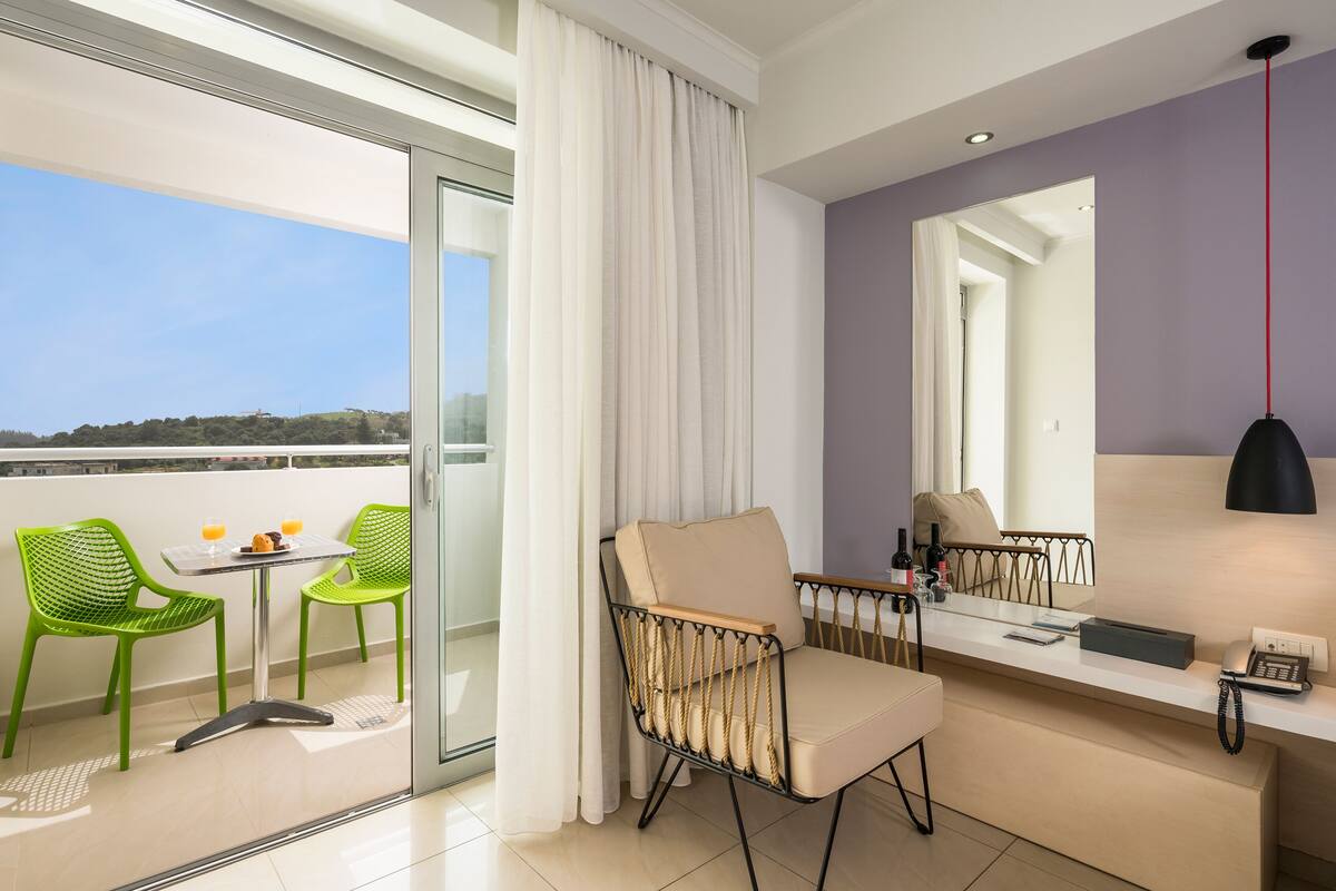 A modern hotel room with a balcony, a sofa, a chair, a small table with a drink, and a mirror.
