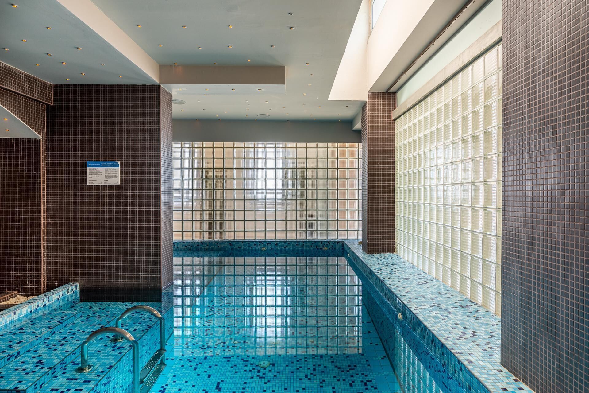 Una piscina interior con paredes y techo azulejados, vista despejada al cielo y un cartel con las normas de la piscina.