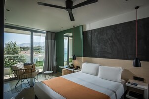 A modern hotel room with a large bed, a desk, a chair, and a balcony with a view of the city and mountains.