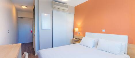 Habitación de hotel con cama, mesita de noche, aire acondicionado de pared y puerta que da a otra habitación.