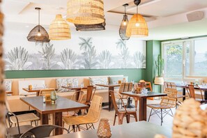 Un restaurant avec des tables et des chaises en bois, des lampes en osier suspendues au plafond et une fresque représentant des plantes tropicales sur le mur.