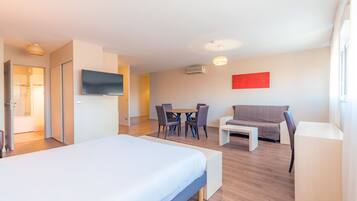 A hotel room with a bed, a TV, a dining table with chairs, a sofa, and an air conditioning unit.