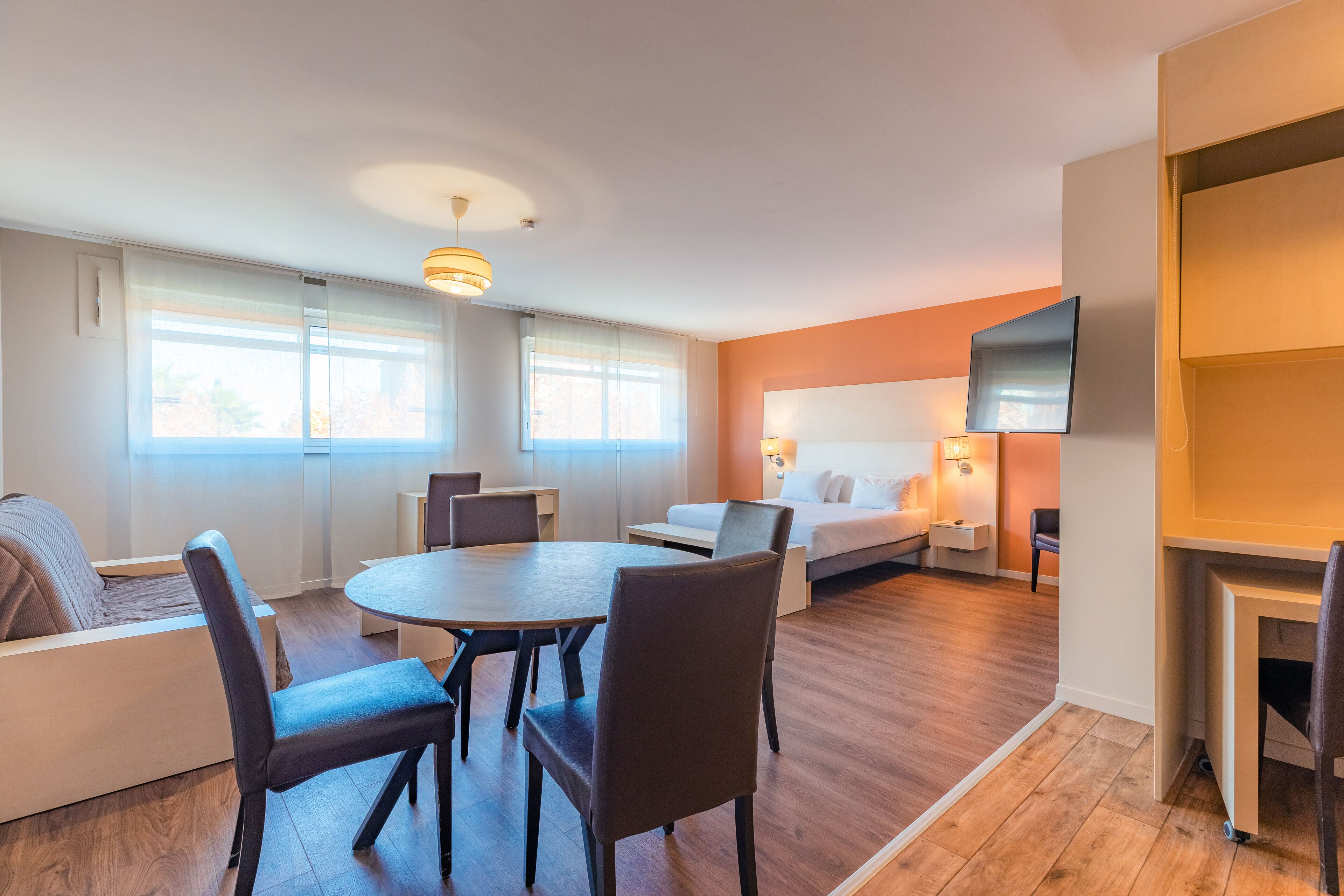 A hotel room with a bed, a TV, a dining table with chairs, a sofa, and a window with curtains.