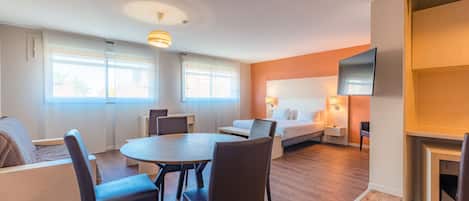 A hotel room with a bed, a TV, a dining table with chairs, a sofa, and a window with curtains.