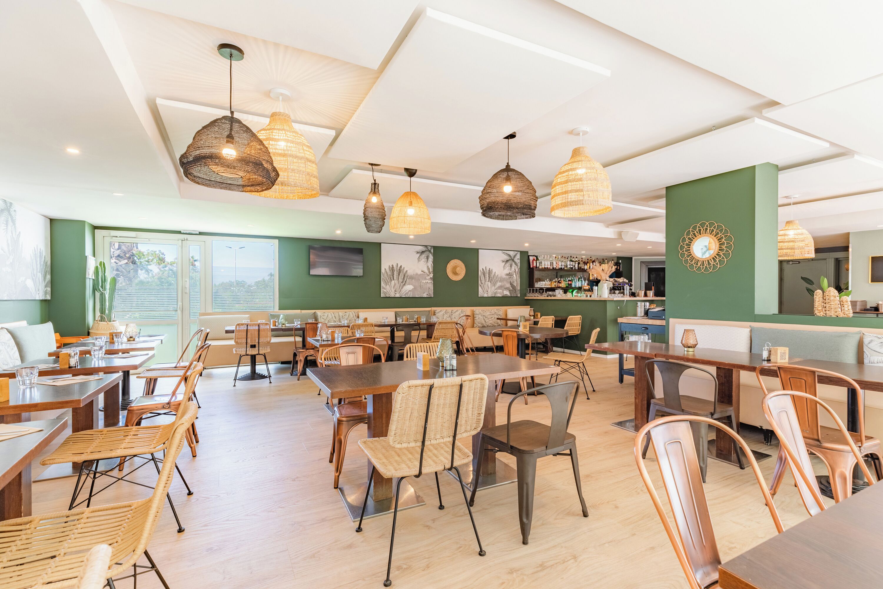 Un restaurante espacioso con varias mesas y sillas, lámparas colgantes de mimbre y una pared de acento verde.