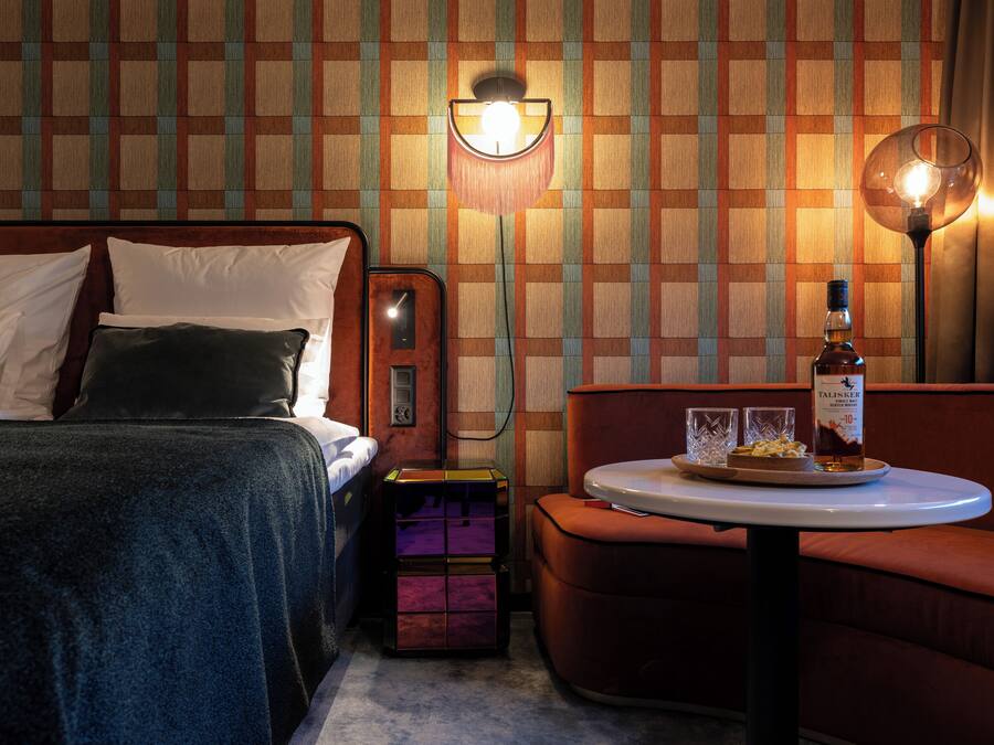 A hotel room with a bed, a small table with a bottle and glasses, and a wall with a checkered pattern.