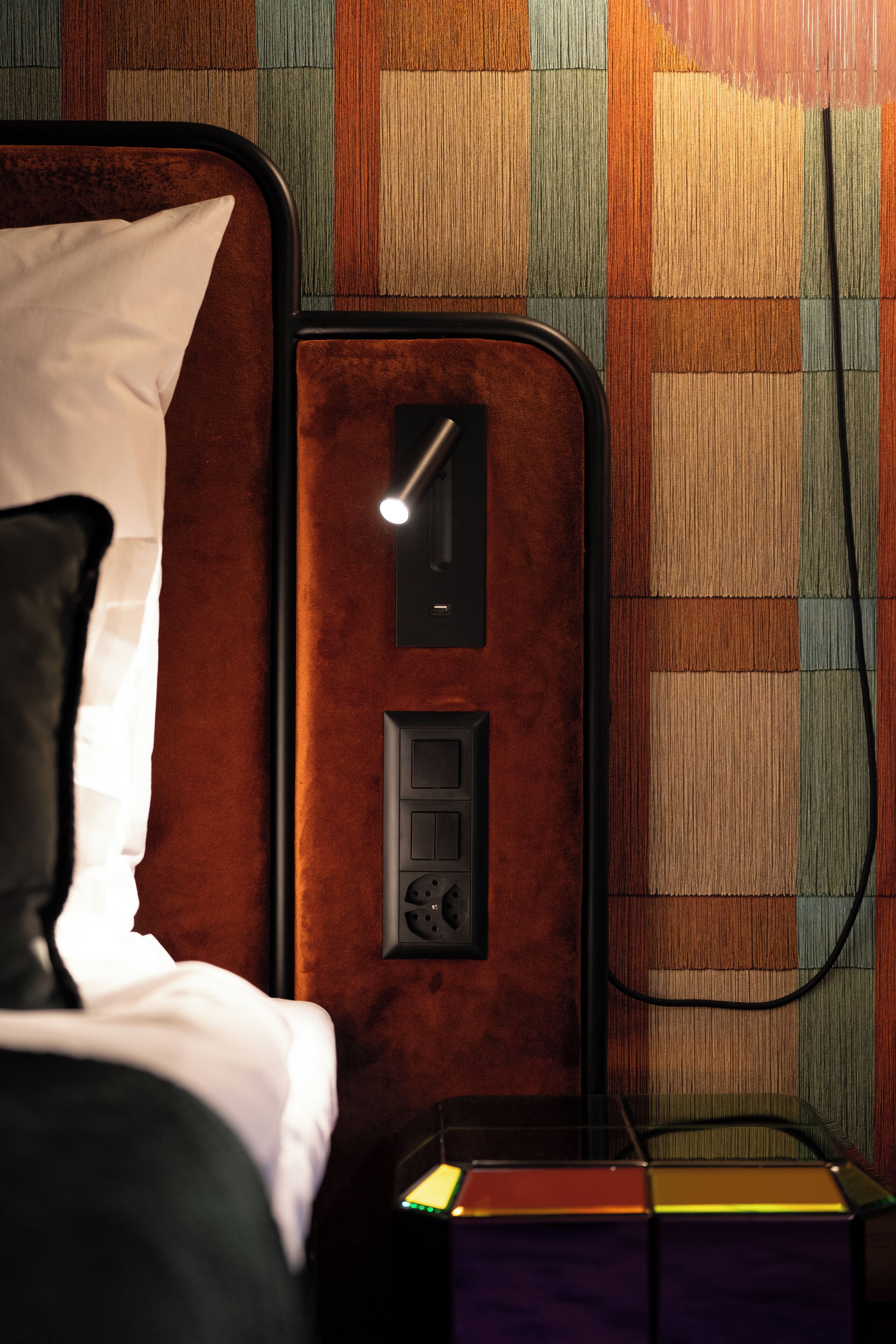 Quarto de hotel com cama, mesa de cabeceira, abajur e parede com padrão listrado.