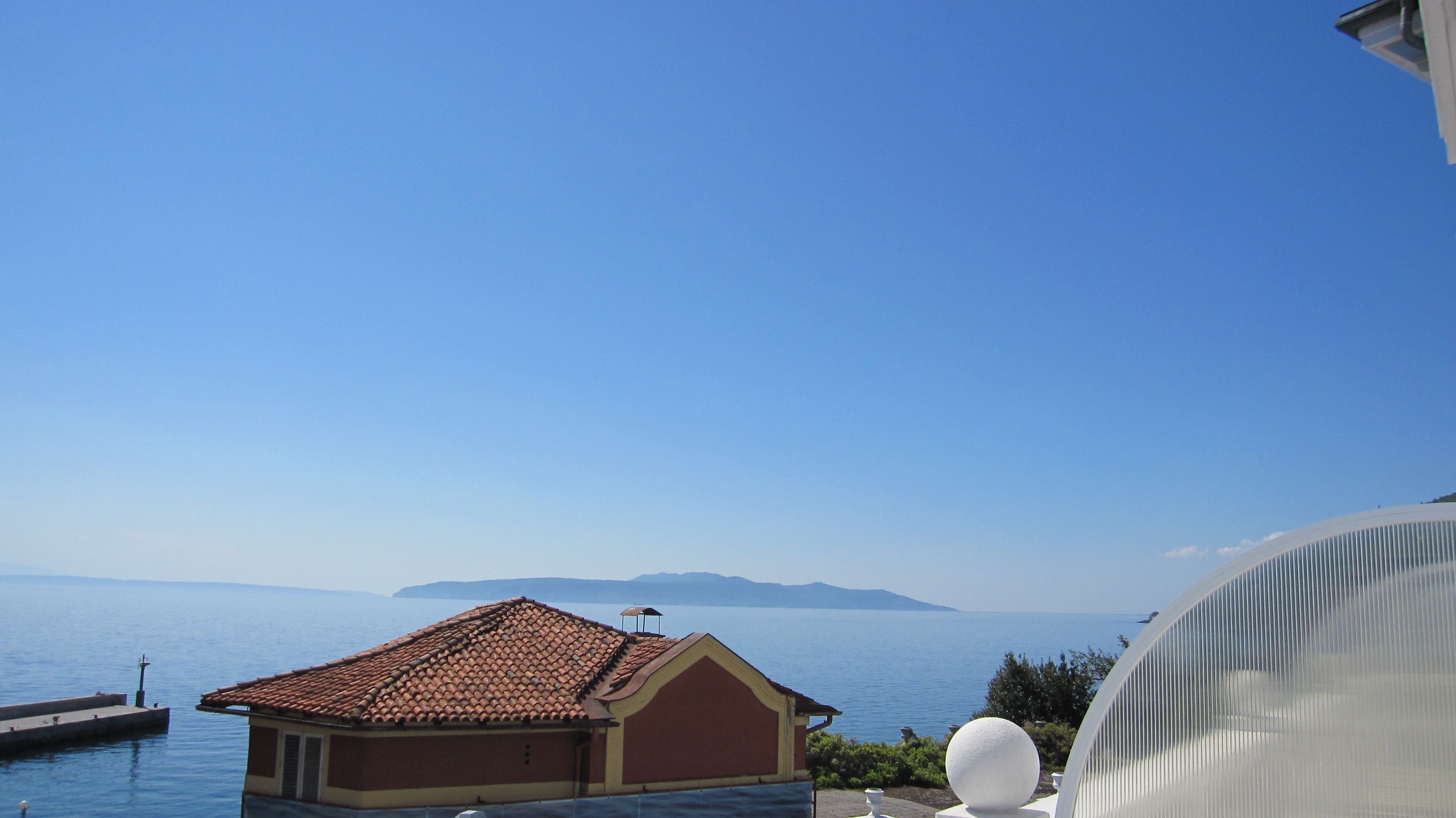 A coastal building with a tiled roof overlooking a calm sea and distant mountains.