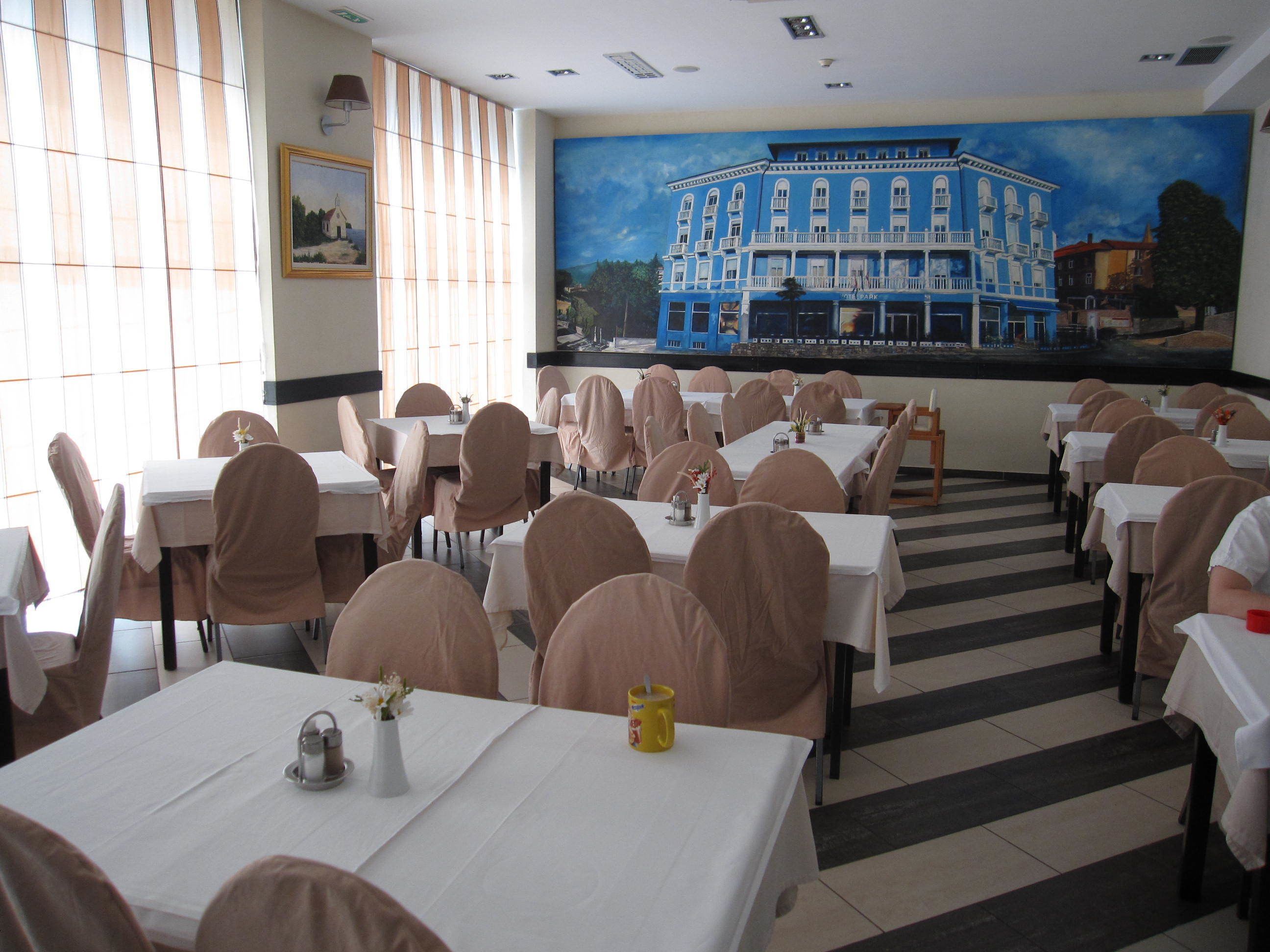 A dining area with tables and chairs, a mural of a building, and a window with blinds.