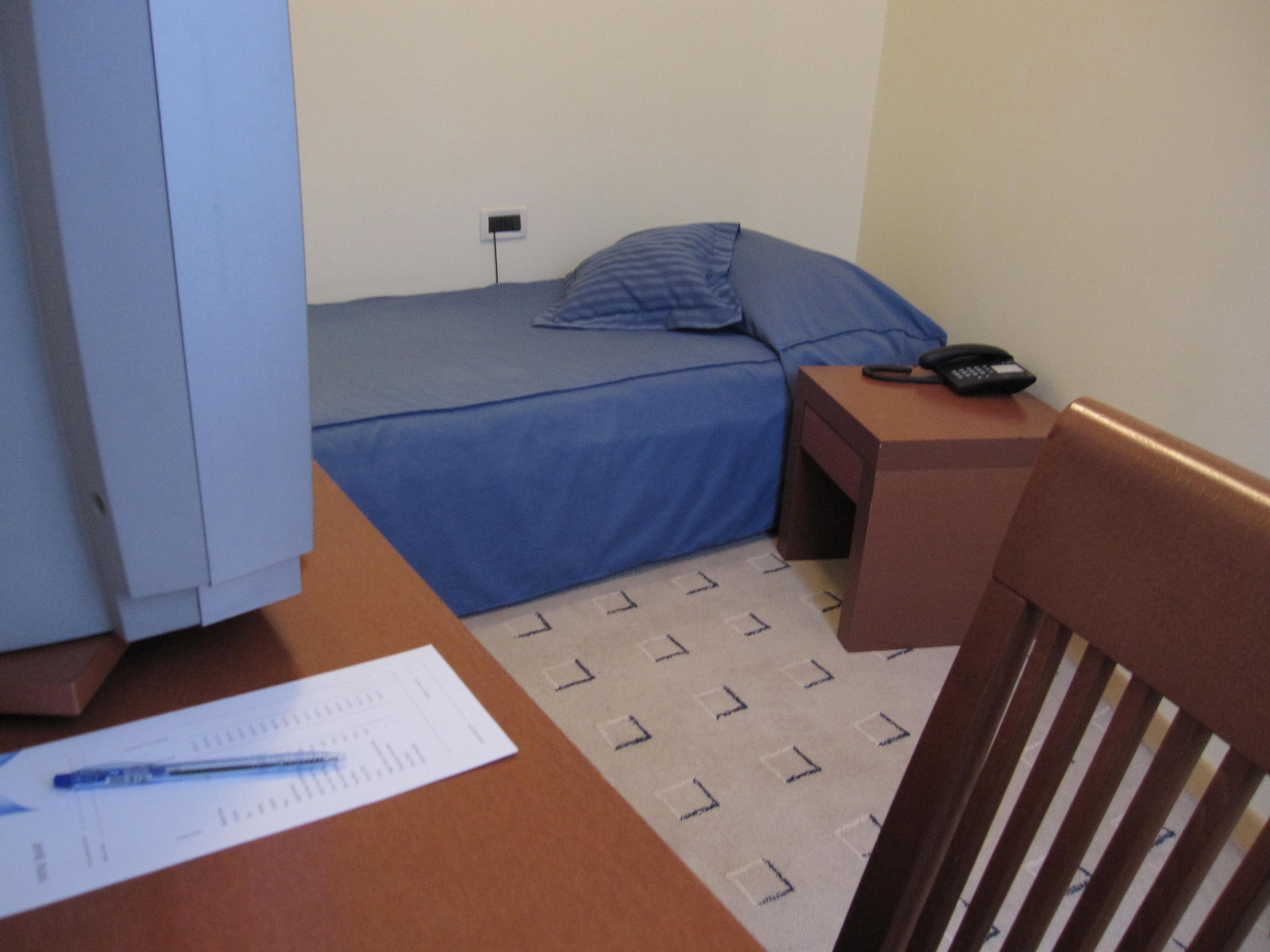 A small, single-bed room with a blue bedspread, a bedside table with a phone, and a chair.