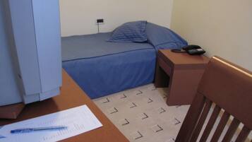 A small, single-bed room with a blue bedspread, a bedside table with a phone, and a chair.