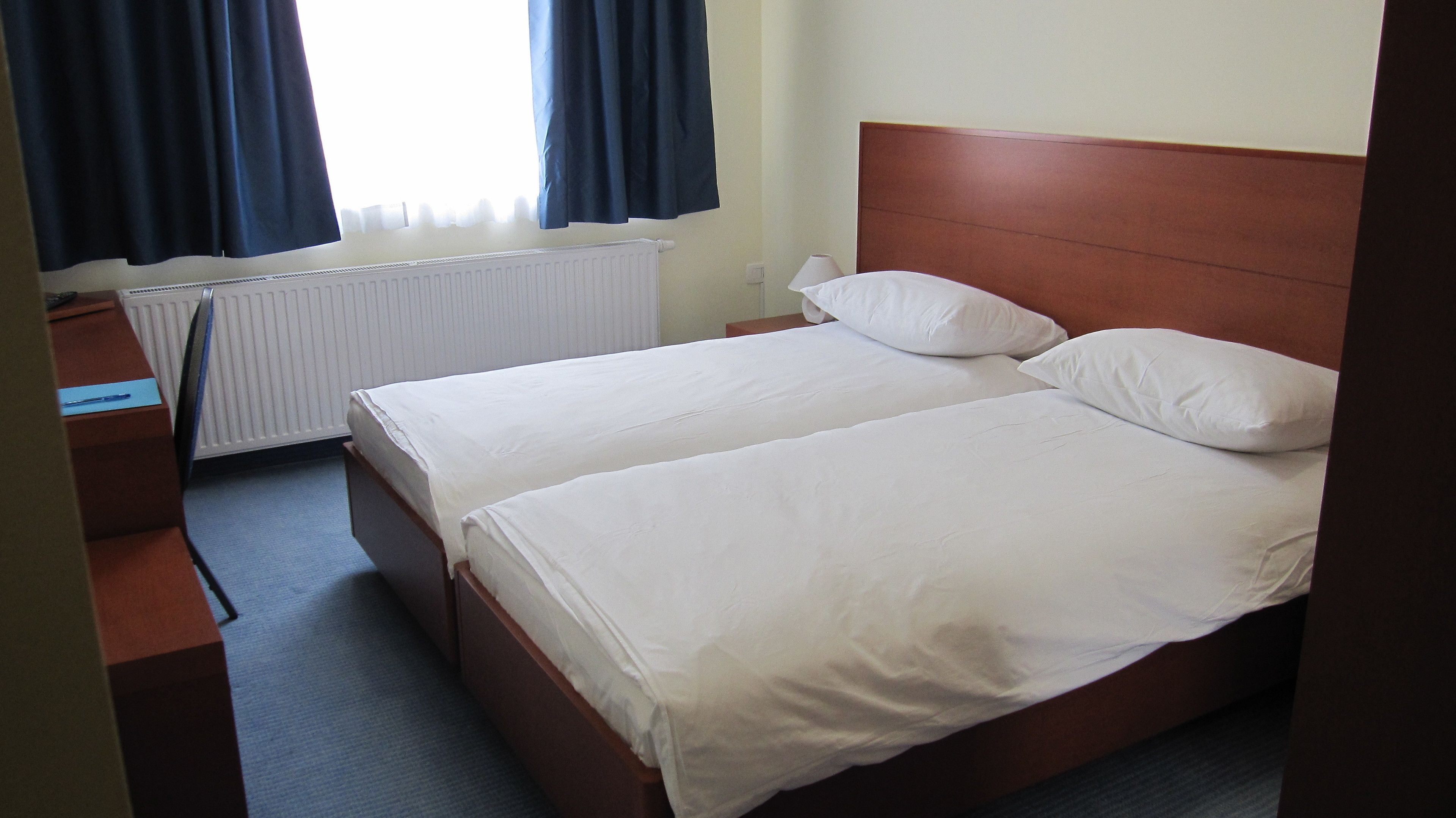 A hotel room with two single beds, a wooden headboard, a desk with a chair, a radiator, and a window with curtains.