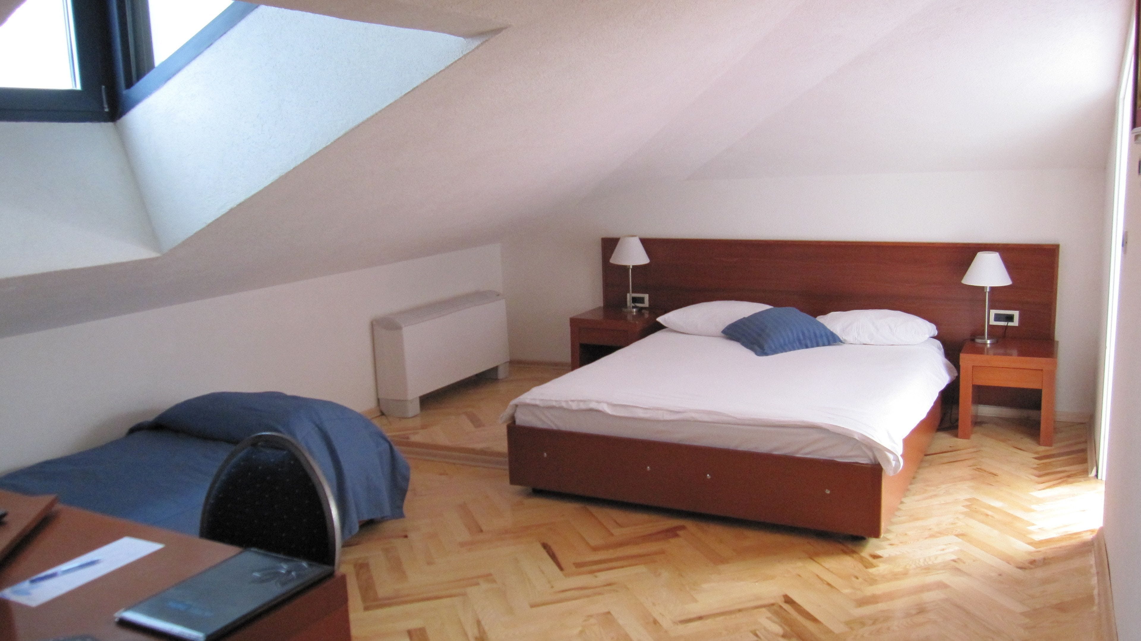 A bedroom with a wooden bed, two bedside tables with lamps, a desk with a chair, and a radiator.