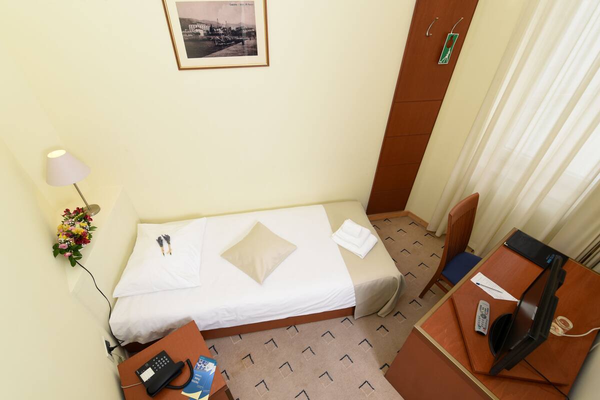A hotel room with a single bed, a desk with a computer, a telephone, and a small table with a remote control.