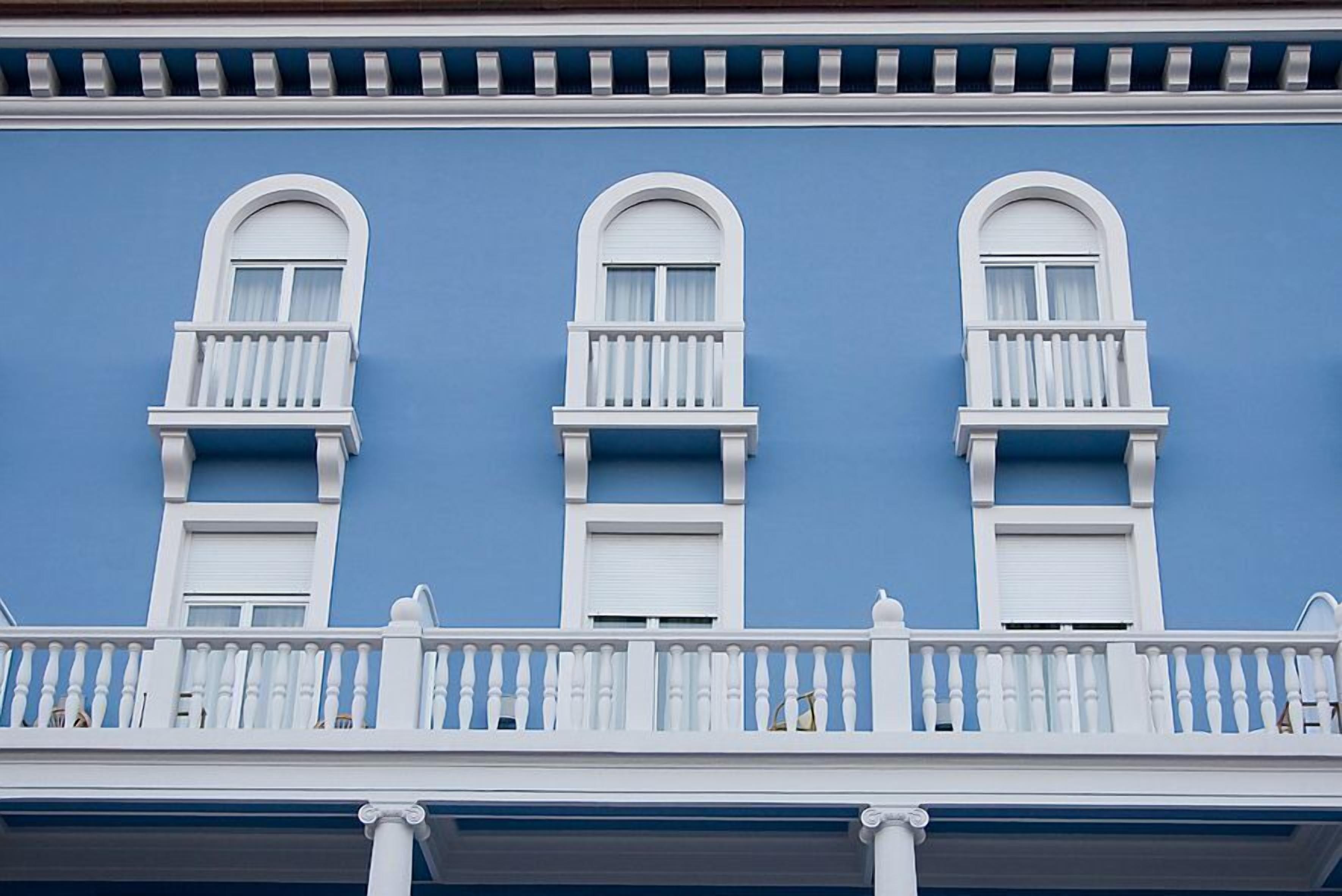 Un bâtiment doté de trois fenêtres à encadrement blanc, chacune surmontée d’un balcon, d’une façade bleue et d’une balustrade de balcon blanche.