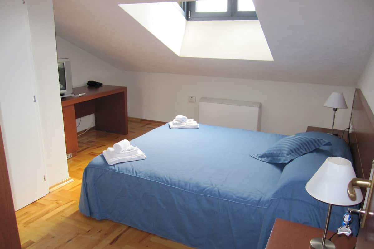 A bedroom with a skylight, a bed with blue bedding, a desk with a computer, and a bedside lamp.