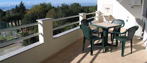 A balcony with a table and chairs, a white railing, and a view of trees and mountains.