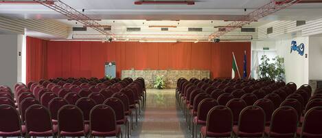 Uma sala de conferências com fileiras de cadeiras vermelhas, palco com um púlpito e um pano de fundo de cortina vermelha.