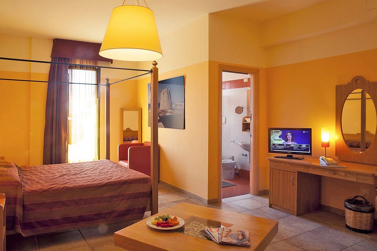 Une chambre d’hôtel avec un lit à baldaquin, une télévision, un bureau avec un ordinateur et une salle de bain visible à travers une porte ouverte.