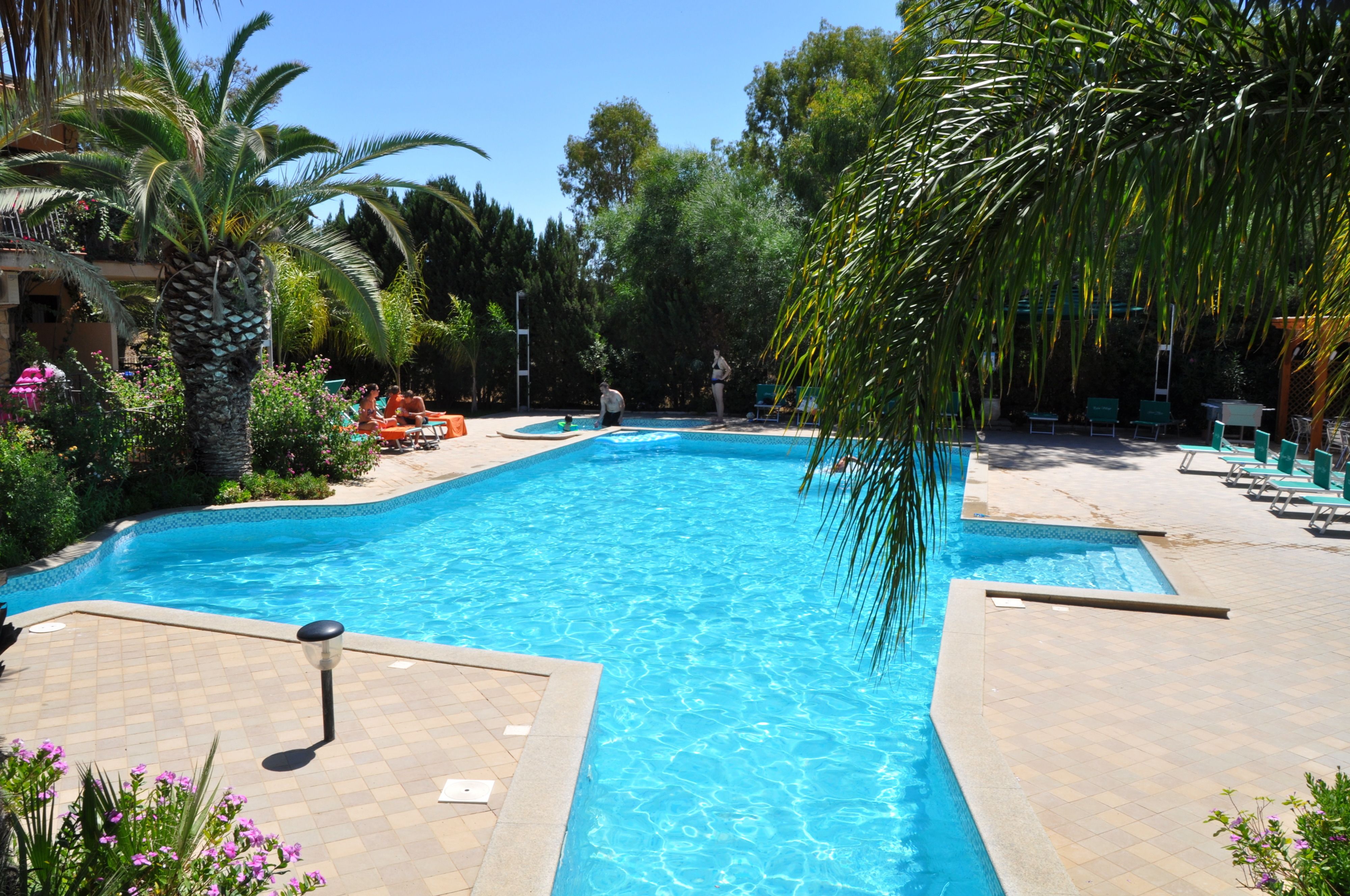 Ein Swimmingpool, umgeben von Palmen und Grünpflanzen, mit Liegestühlen und einer gefliesten Liegefläche.
