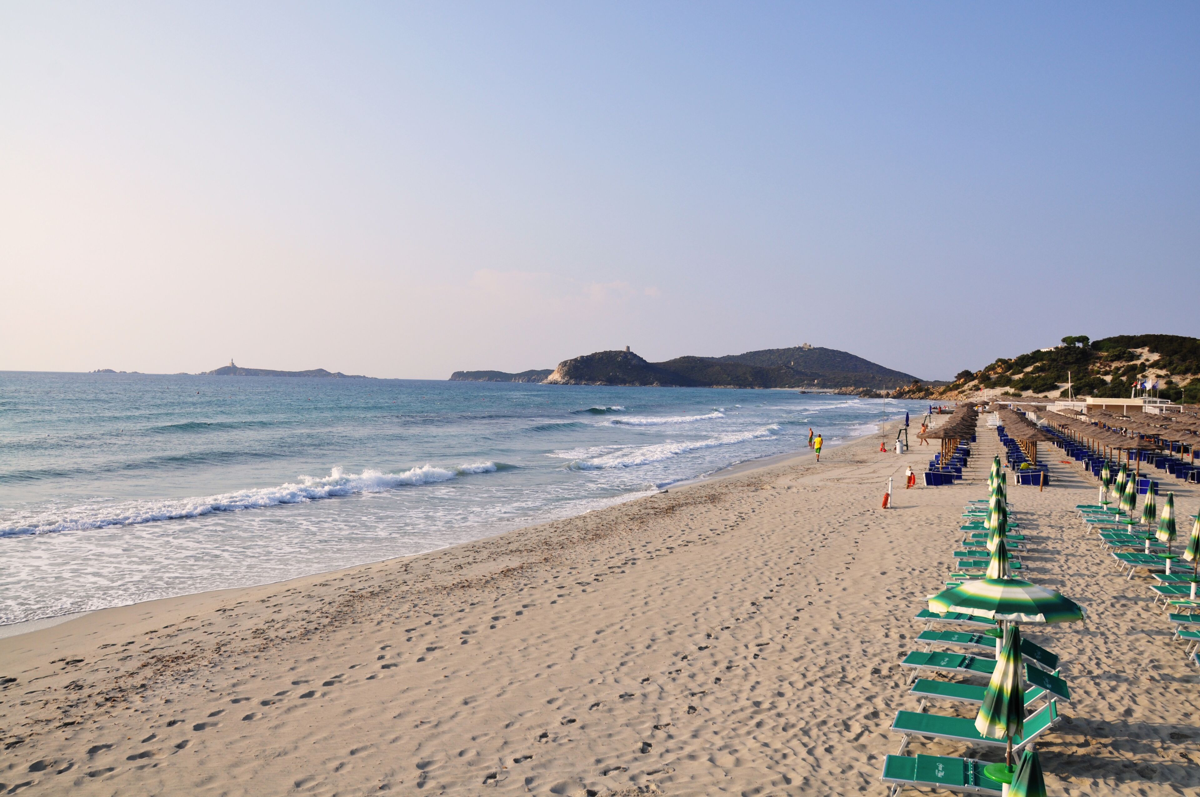A beach with rows of sun loungers and umbrellas, a clear sky, and a distant hill with a lighthouse.