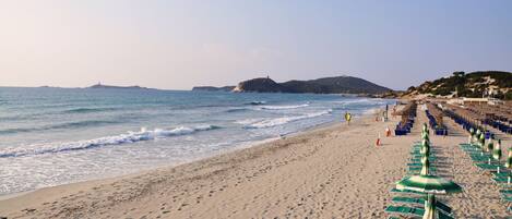 A beach with rows of sun loungers and umbrellas, a clear sky, and a distant hill with a lighthouse.