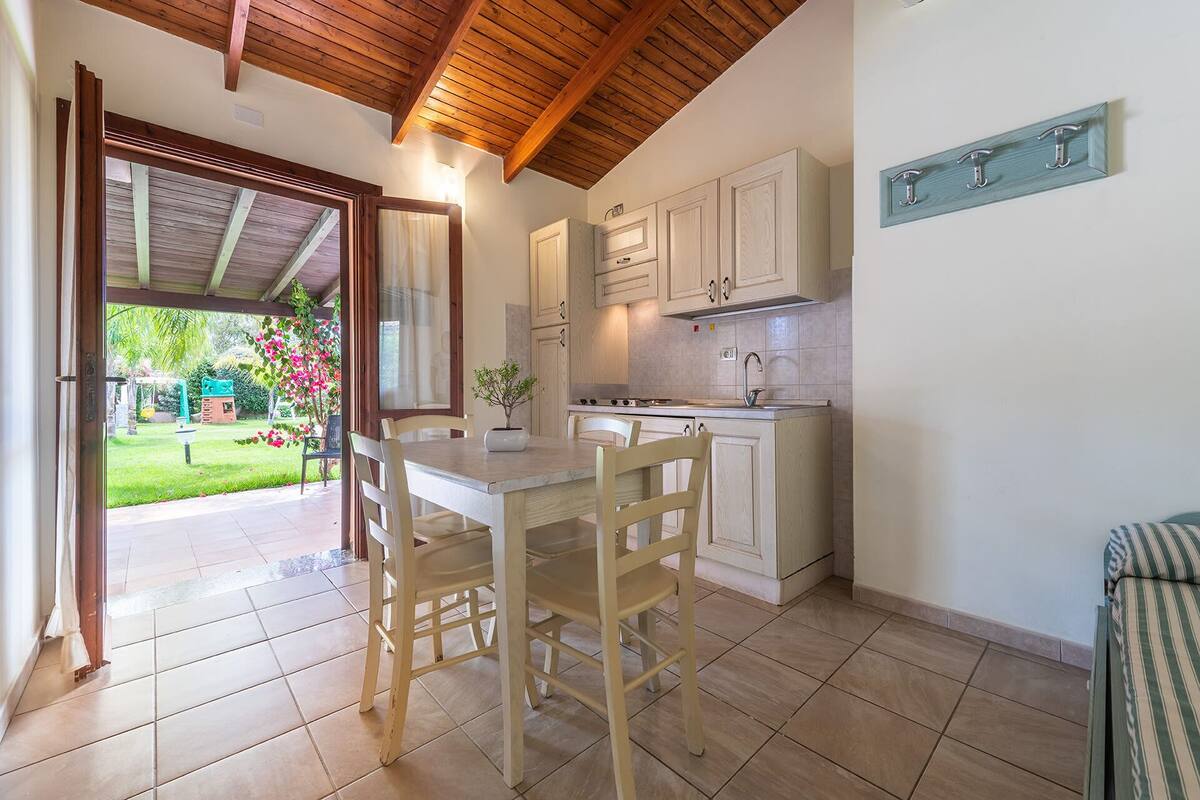 A kitchen with wooden cabinets, a dining table with chairs, and a view of a garden through an open door.
