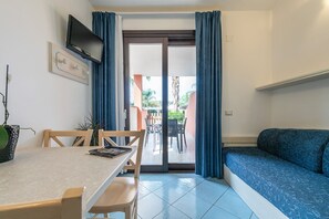 A room with a dining table, chairs, a sofa, a TV mounted on the wall, and a sliding glass door leading to an outdoor area with a palm tree and a patio table. - Green Village Resort (Villasimius)