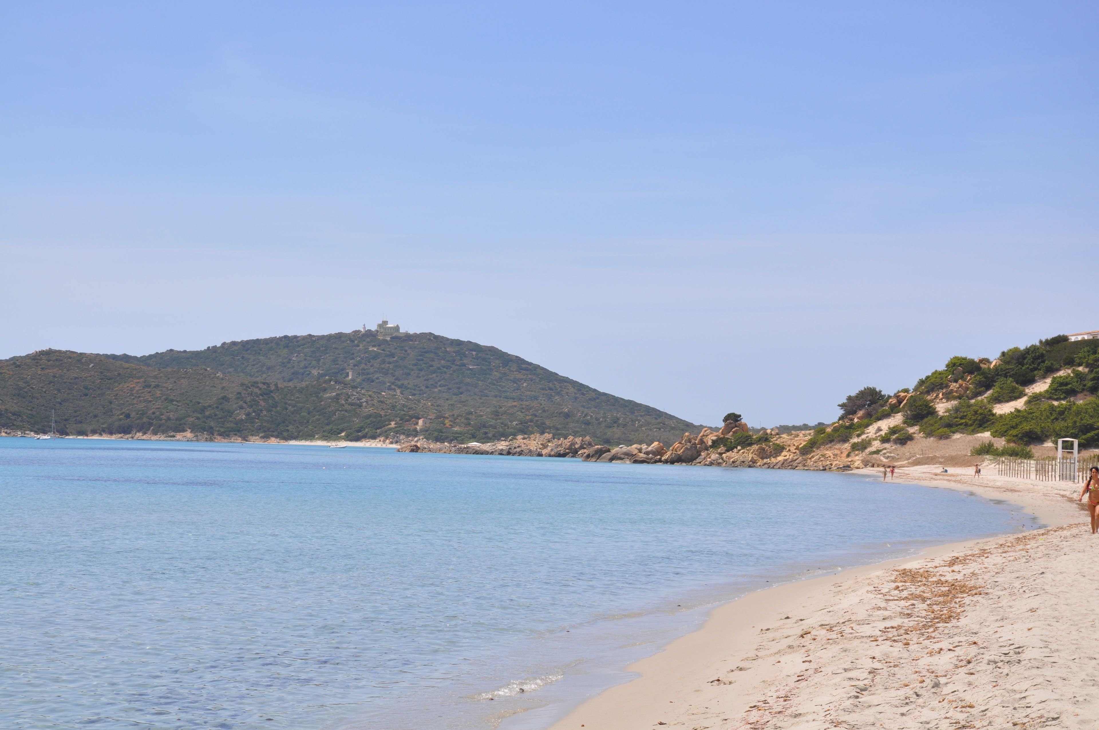 A beach with clear blue water, sandy shore, and a hill with a structure on top.