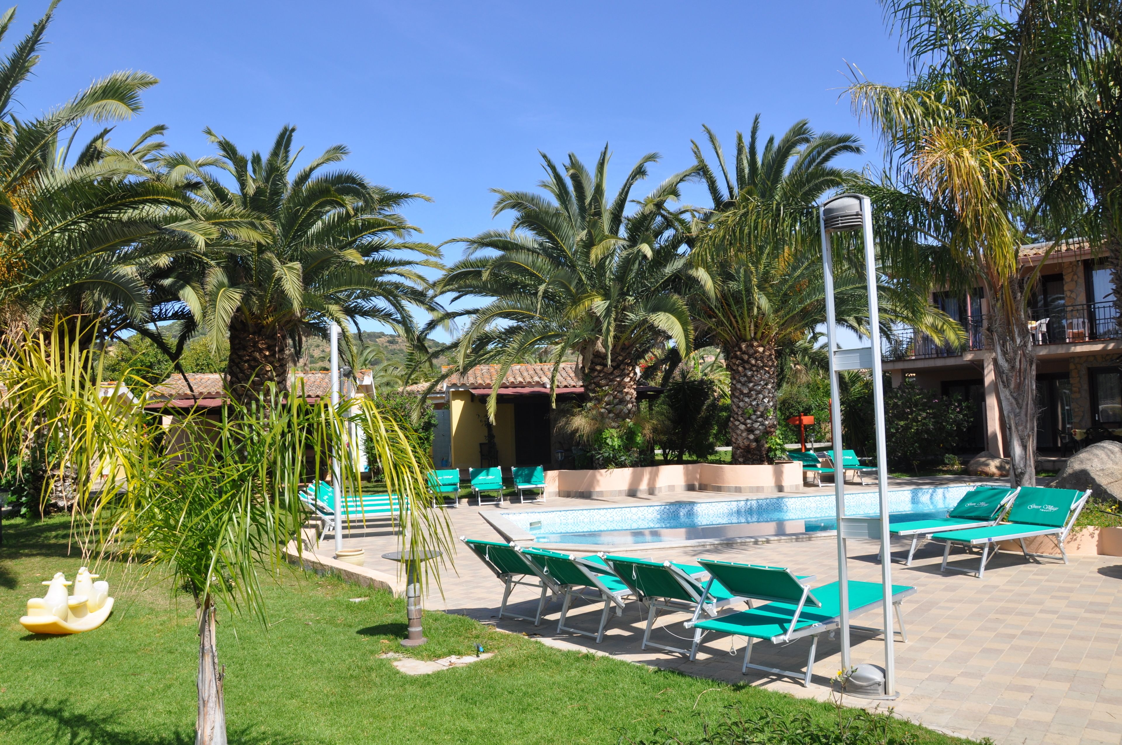 Zona de piscina con sillas de descanso verdes, palmeras y cielo azul despejado.