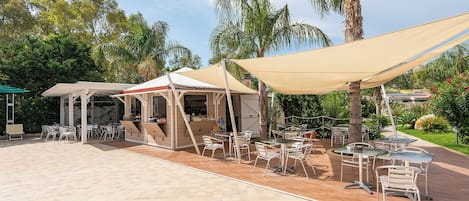 An outdoor patio area with multiple tables and chairs under large umbrellas, surrounded by palm trees and greenery.