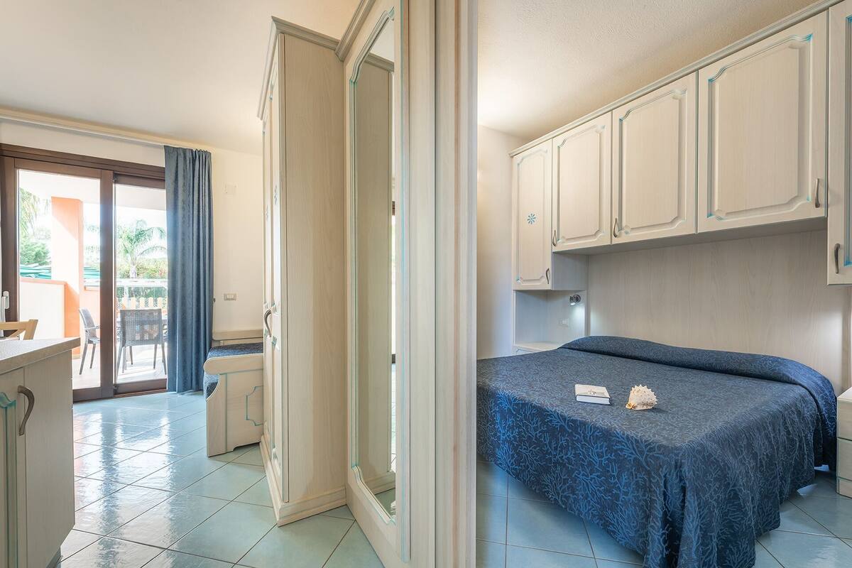 A bedroom with a bed, a small desk, and a chair. There is a sliding door leading to a balcony with a table and chairs.