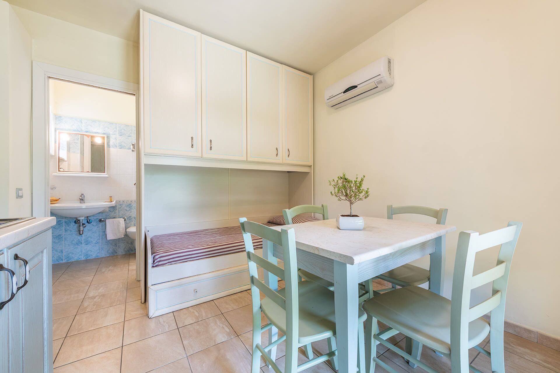 Una cocina compacta con armarios blancos, una mesa de comedor con sillas, un lavamanos pequeño y una planta en maceta.