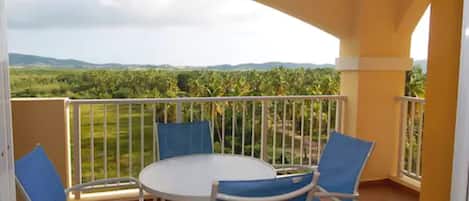 A balcony with a white table and blue chairs, offering a view of a green landscape with palm trees.
