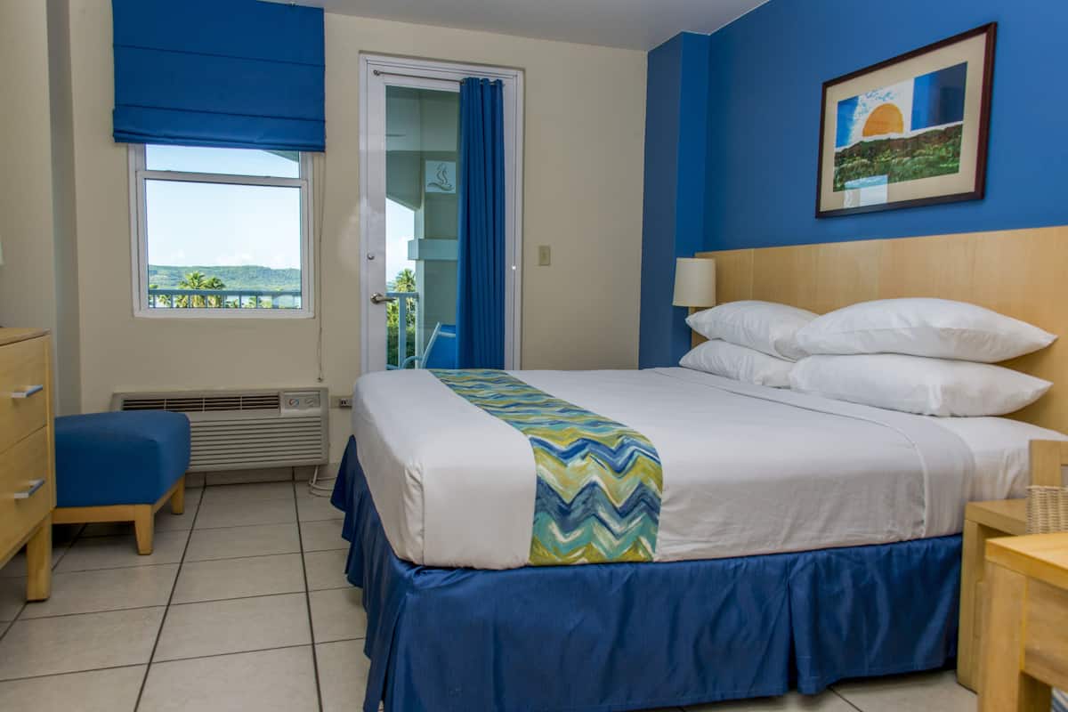 A hotel room with a bed, a desk, a chair, a window with a view, and a painting on the wall.