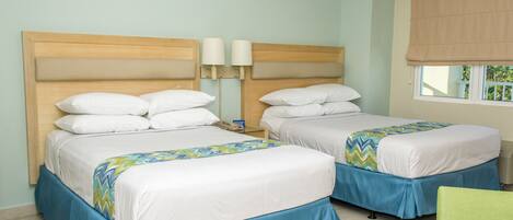 Habitación de hotel con dos camas, cada una con cabecero de madera, ropa de cama blanca y faldas azules. Hay dos lámparas de noche y una ventana con vistas a zonas verdes.