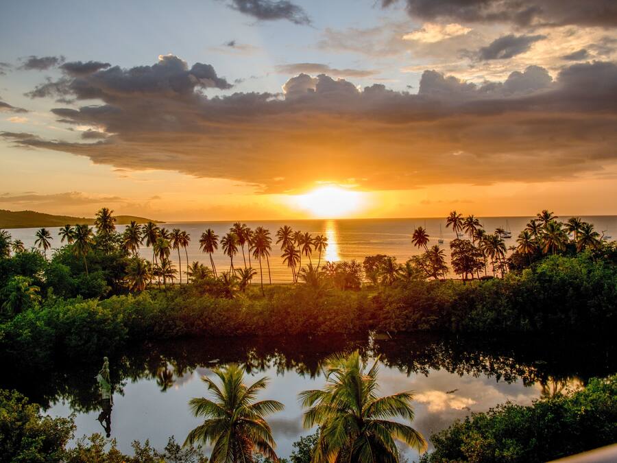 A sunset over a calm body of water with palm trees and a distant shoreline.