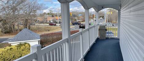 A covered porch with white railings and a blue floor, overlooking a street with cars and a building in the background.